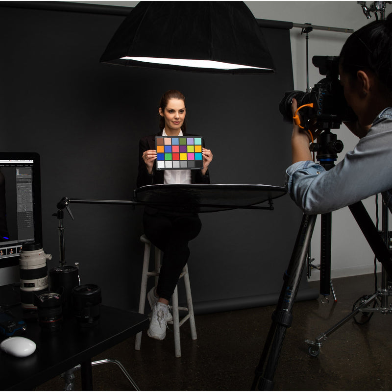 Een afbeelding van de Calibrite ColorChecker Classic die door het onderwerp in een portret tijdens een fotoshoot wordt vastgehouden. De kleurreferentiekaart kan door fotografen worden gebruikt als referentie om te voorspellen en te controleren hoe kleur eruit zal zien onder verschillende lichtomstandigheden en om een nauwkeurige kleurweergave tijdens het bewerken te waarborgen. Het toont 24 vierkanten, elk met een kleurreferentie.